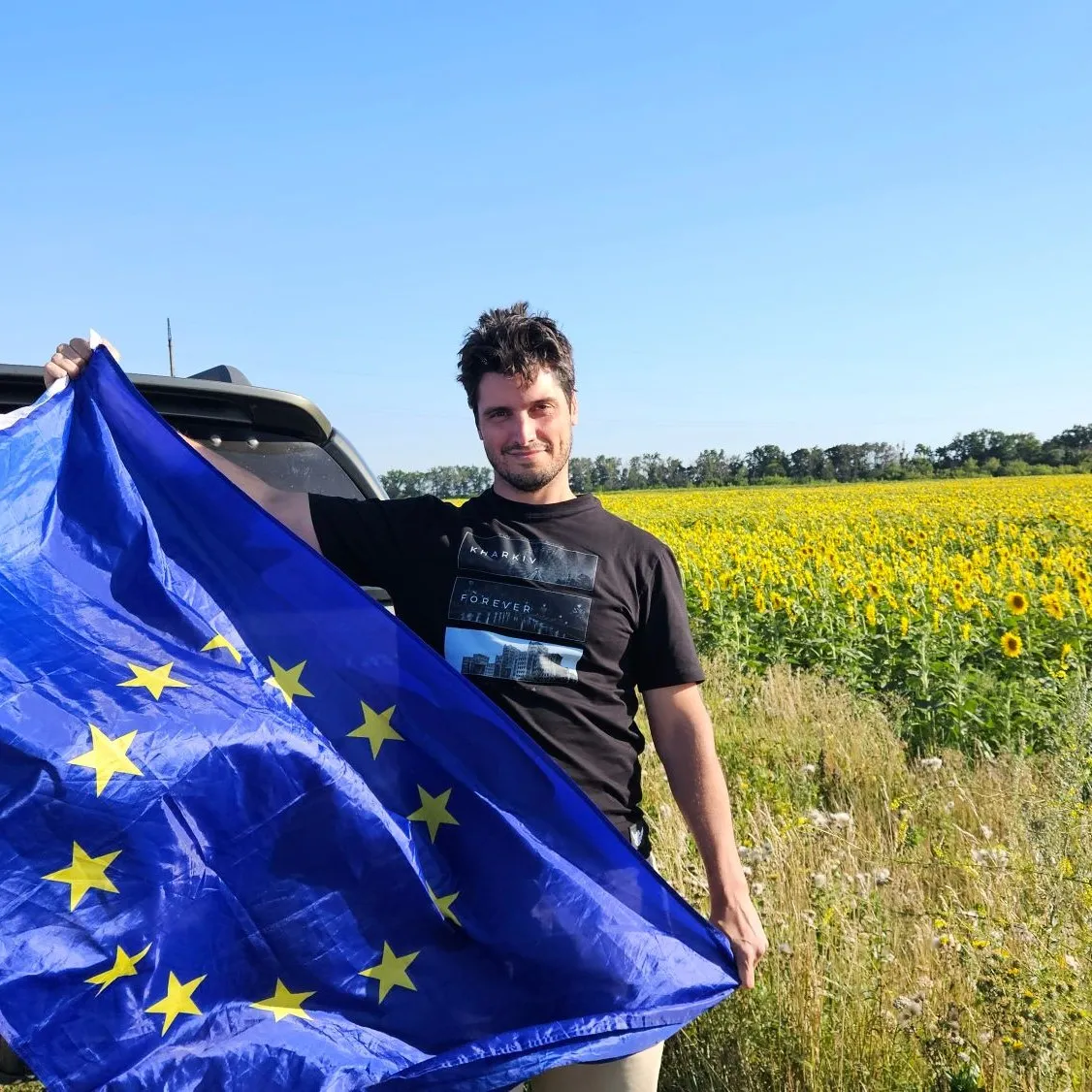 Carlos holding a European Union flag beside the donated truck and a sunflower field in the Kharkiv region